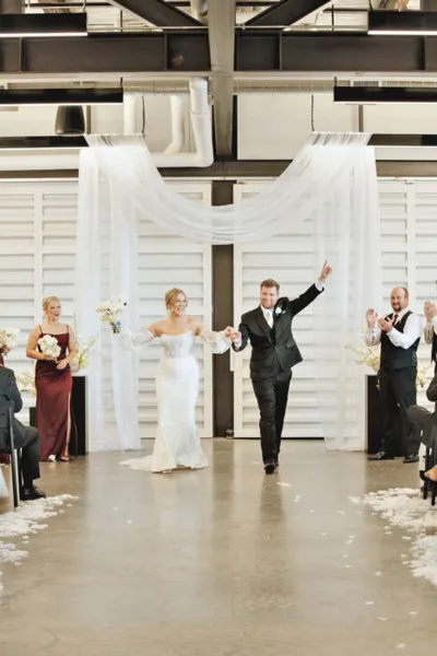 Bride and groom walk jubilantly back down the aisle in an industrial-style venue with white draping