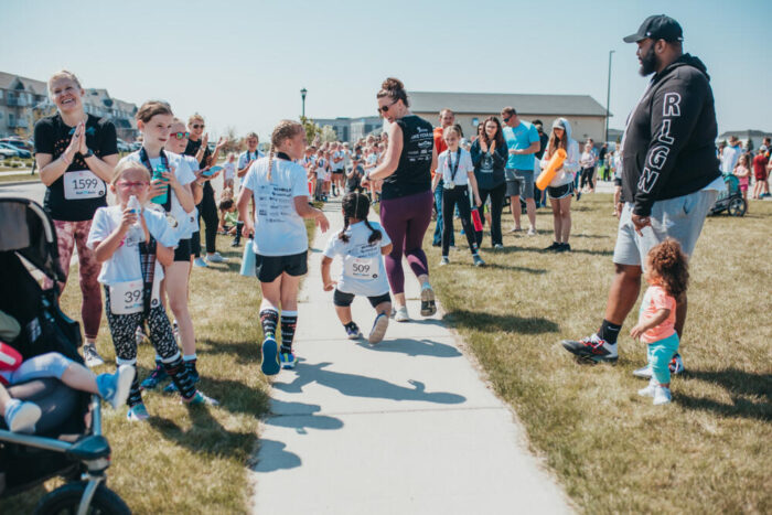 Children and adults in race bibs celebrate along a sunny outdoor fun run course