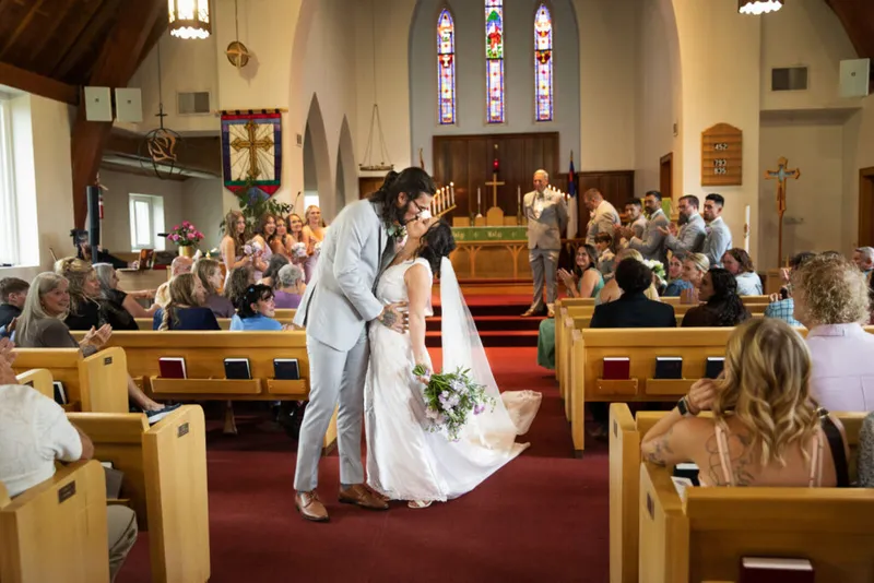 Bride and groom share a kiss in the aisle of a church while guests cheer from wooden pews