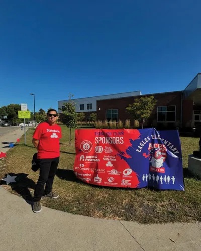 Man in red State Farm shirt standing beside Eagles Elementary sponsors banner outdoors