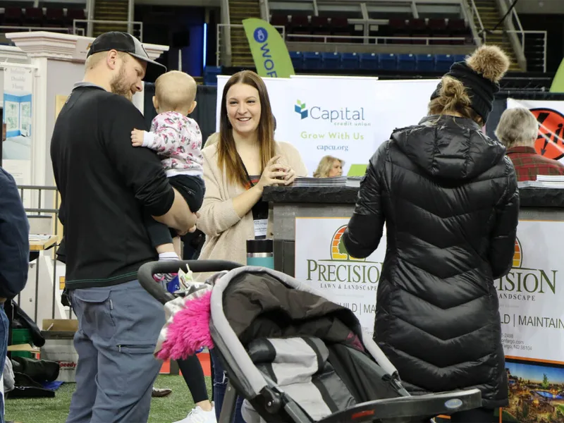 Man holding a baby and a woman chat with a vendor at an indoor home show expo