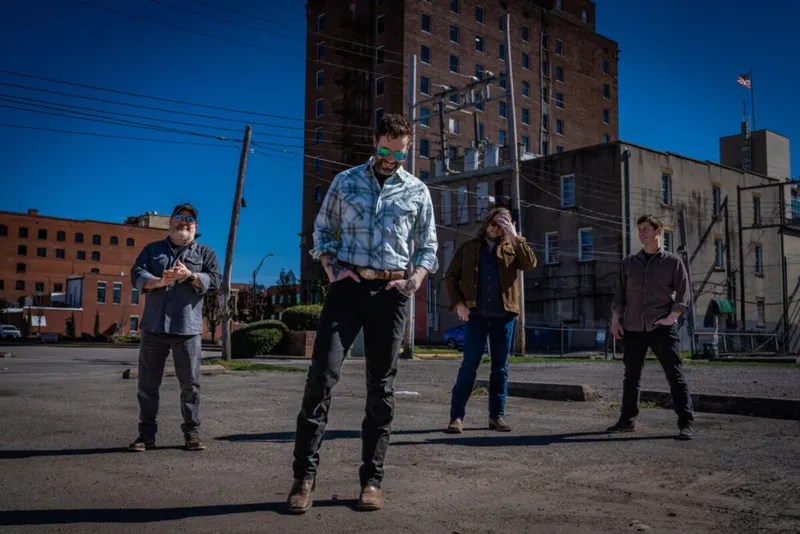 Four men in casual western attire pose in a sunny urban parking lot beside a tall brick building