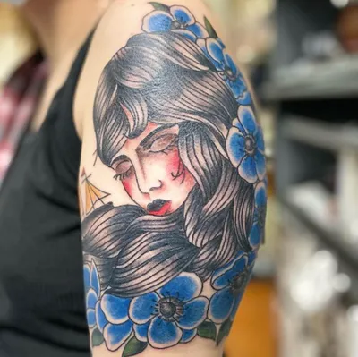 Traditional-style tattoo of a woman with flowing hair surrounded by blue flowers on an upper arm