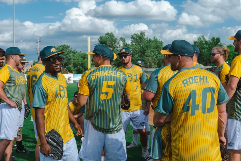 Softball players in yellow and teal AO1 jerseys huddle on a sunny field