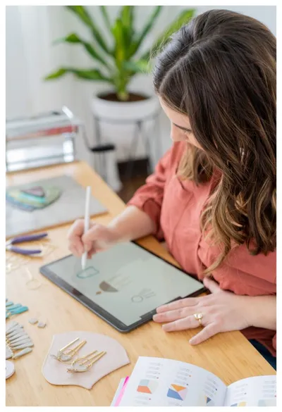 Woman with stylus drawing on a tablet at a wood desk scattered with handmade earrings and a color swatch book