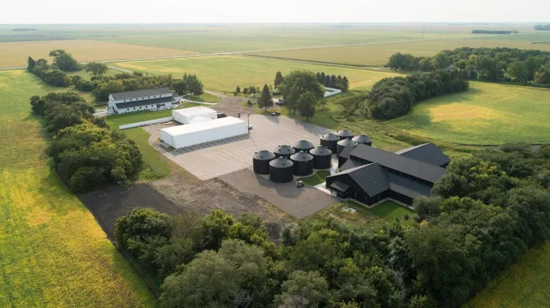 Aerial view of a rural wedding venue with black grain bins, white tent, and dark-roofed barn amid farm fields