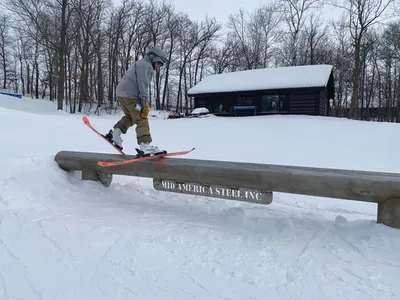 Skier grinding a snow-covered rail at a ski hill, log cabin visible behind