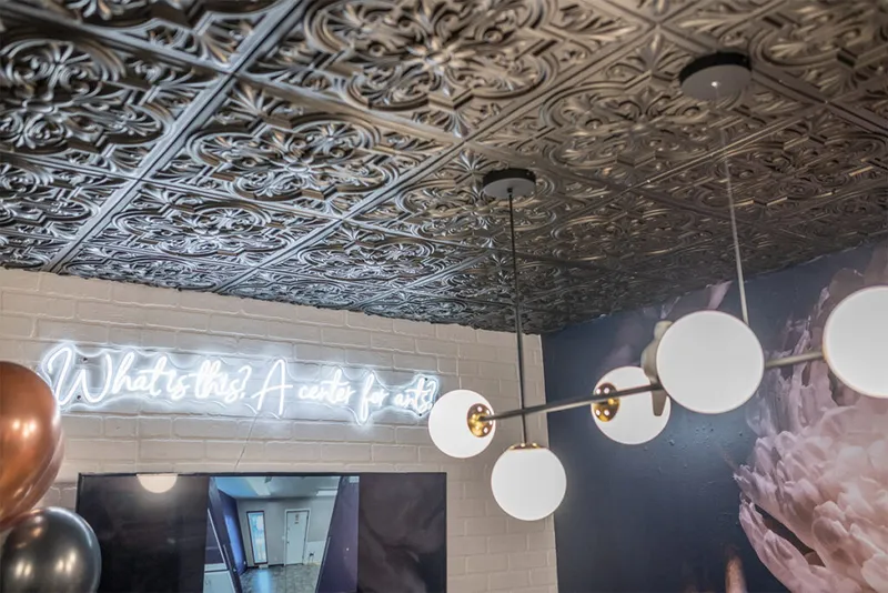 Ornate dark pressed-tin ceiling above a neon sign reading 'What is this? A center for arts' with globe pendant lights