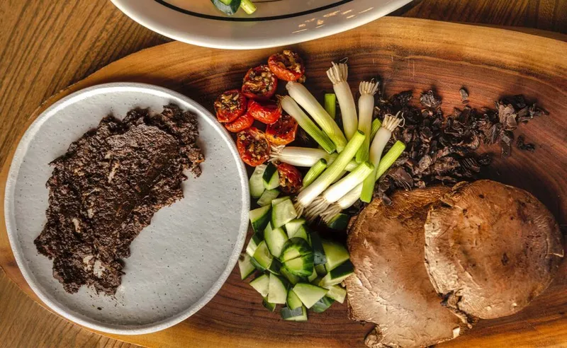 Wooden board with sliced mushroom, scallions, cucumber, dried tomatoes, and dark paste in a ceramic bowl