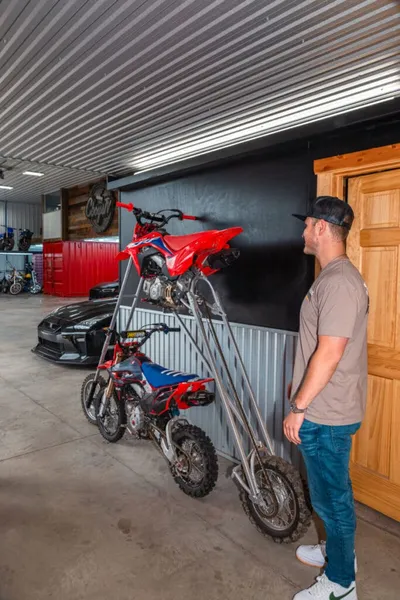 Man in jeans and cap standing beside two dirt bikes stacked on a wall-mounted rack in a garage