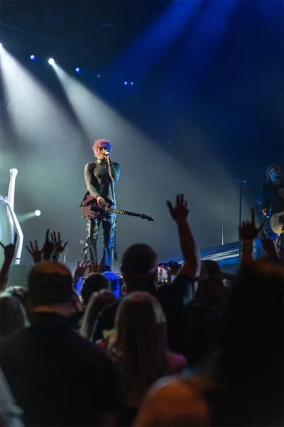 Pink-haired guitarist singing into a mic on a blue-lit stage before a crowd with raised hands