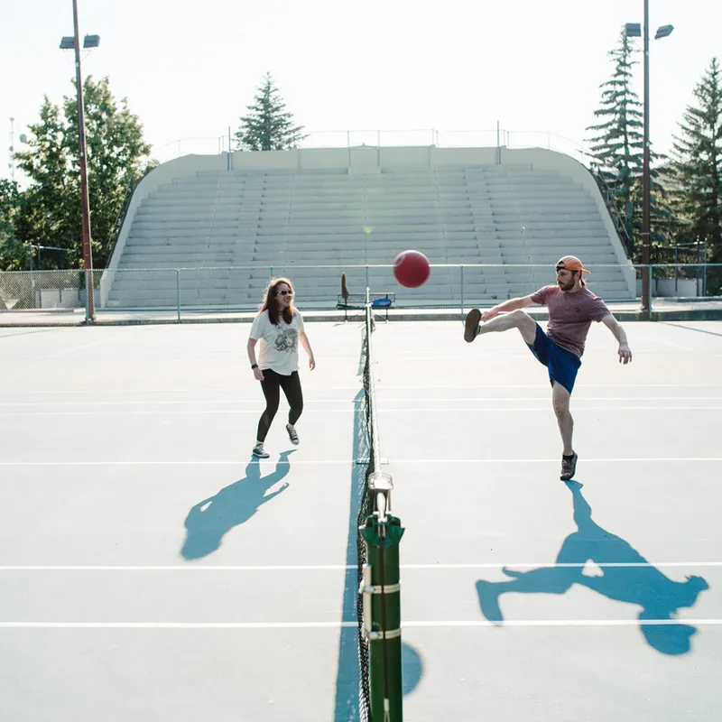 Two people kicking a red ball over a tennis net on an outdoor court with bleachers behind them