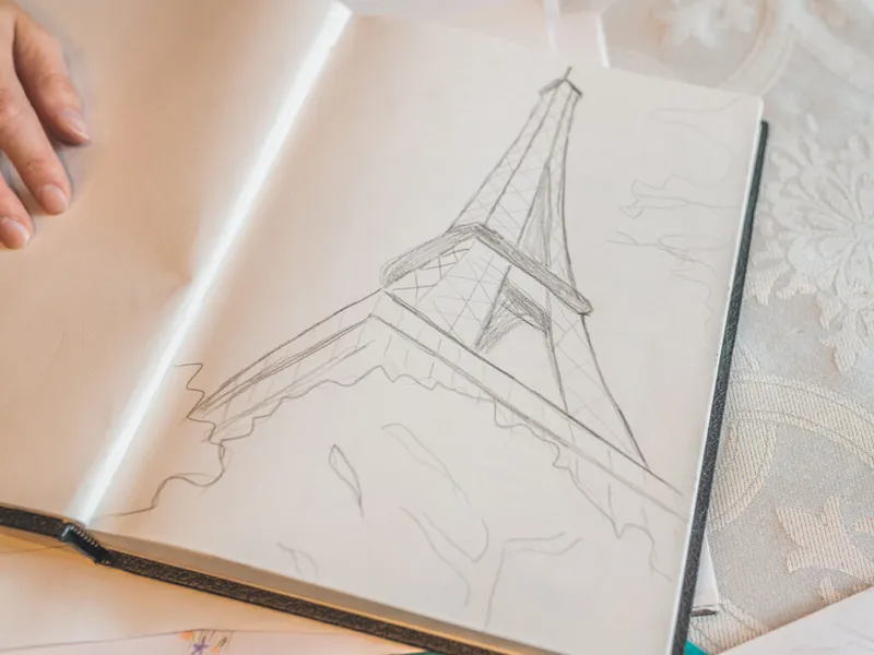 Hand holding open a sketchbook with a pencil drawing of the Eiffel Tower