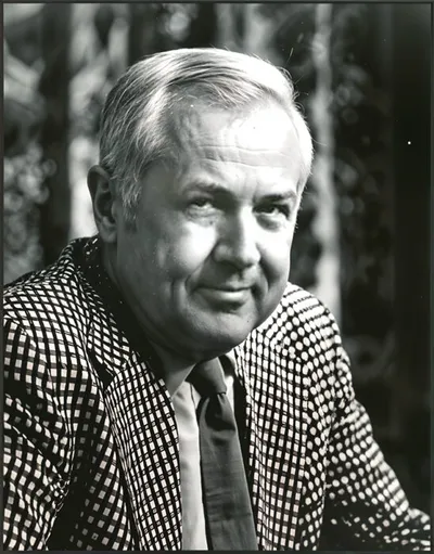 Older man with silver hair smiling, wearing a checkered blazer and tie, black-and-white portrait photo