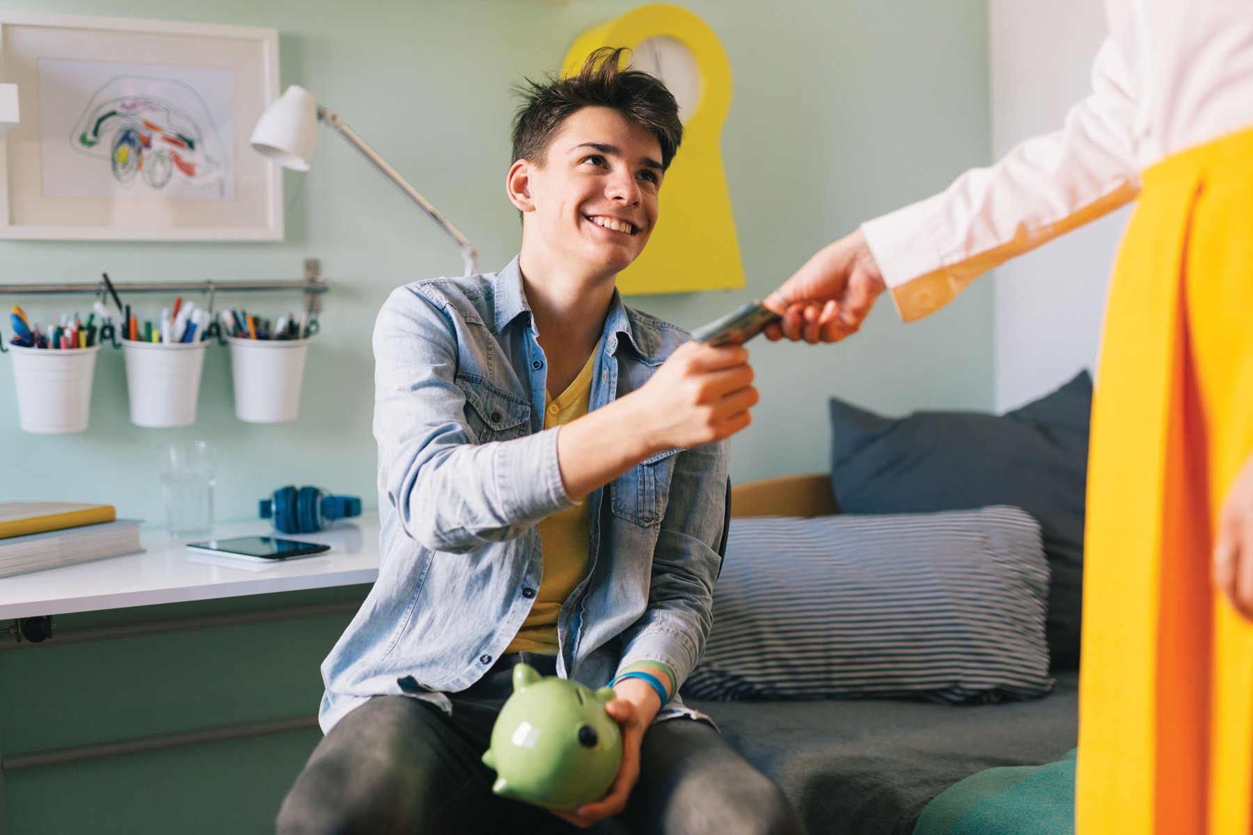 A smiling teenager holding a green piggy bank accepts cash from a parent