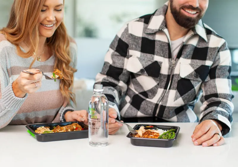 Smiling couple eating prepared meals from black trays at a white counter with a water bottle nearby