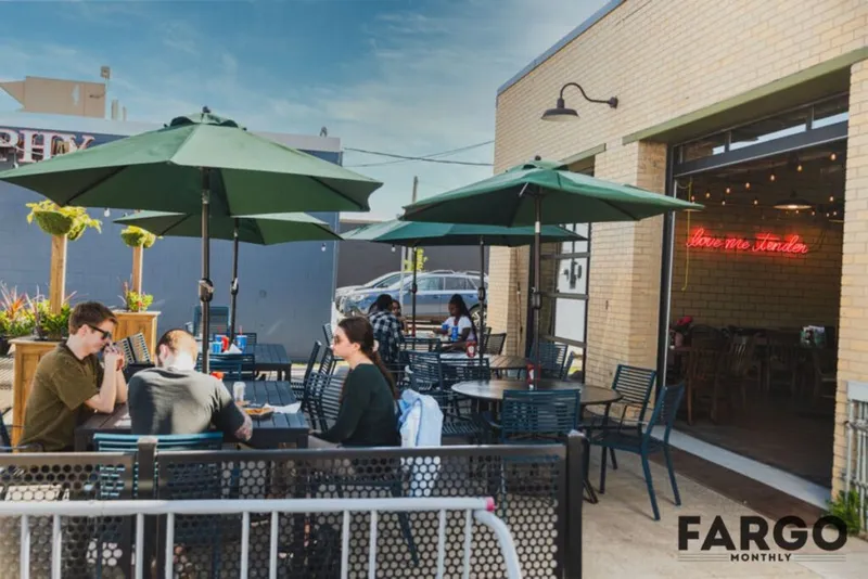 Outdoor restaurant patio with green umbrellas and a red neon 'love me tender' sign on a brick wall