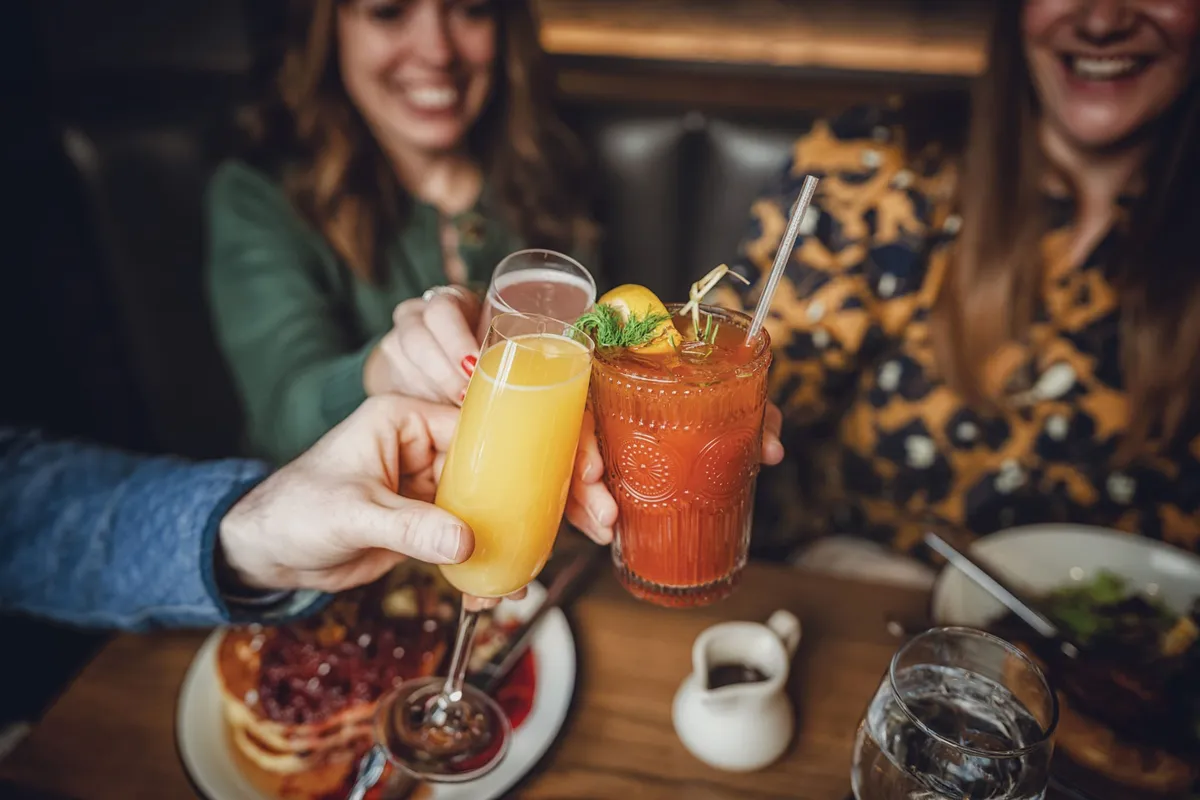 Two smiling women clinking a mimosa and a Bloody Mary over a brunch table