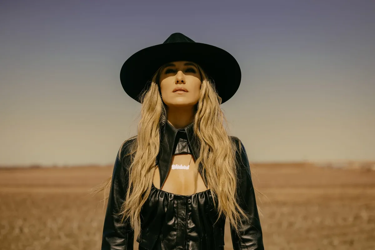 Woman with long blonde hair in black wide-brim hat and leather jacket stands in an open field