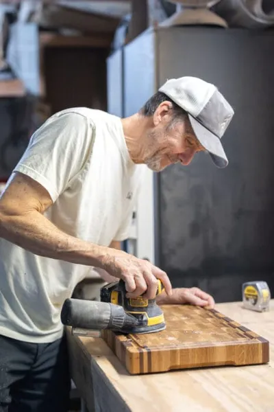 Man in white t-shirt and cap sanding a wooden cutting board in a workshop