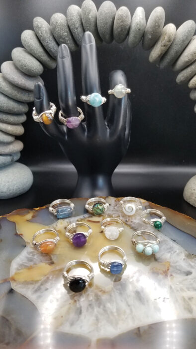 Wire-wrapped gemstone rings displayed on a black hand stand and agate slice, surrounded by smooth stones