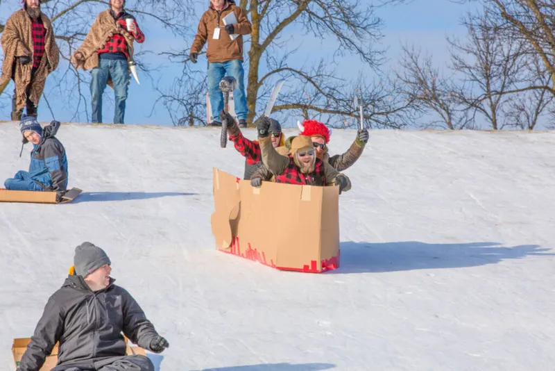 Costumed sledders riding a cardboard box down a snowy hill at a winter community event