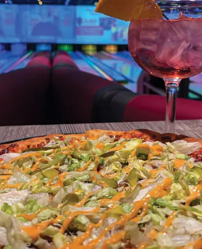 Pizza topped with lettuce, avocado, and drizzled sauce beside a pink cocktail in a bowling alley
