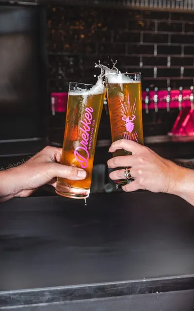 Two hands clinking Drekker Brewing Co. pint glasses filled with golden beer