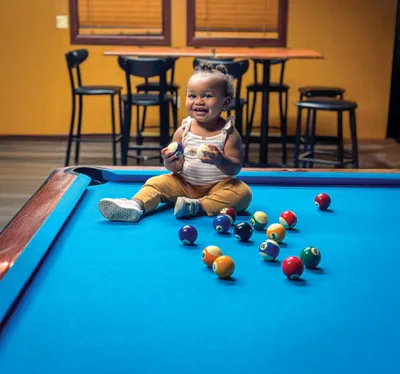 Smiling toddler sitting on a blue pool table surrounded by billiard balls