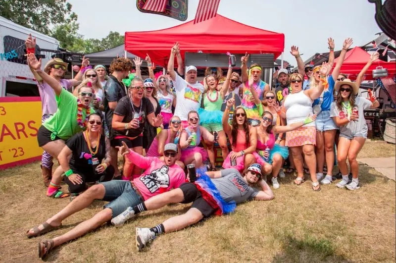 Large group in neon 80s-themed costumes cheering under a red canopy at an outdoor festival