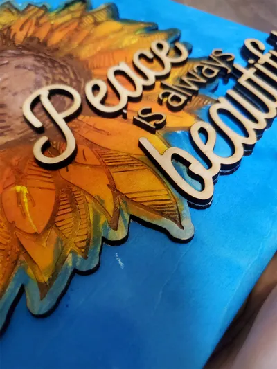 Wood-cut quote "Peace is always beautiful" layered over a sunflower resin art panel