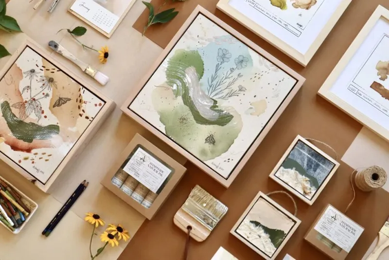 Flatlay of botanical watercolor art prints, kraft boxes, pencils, and yellow flowers on tan background