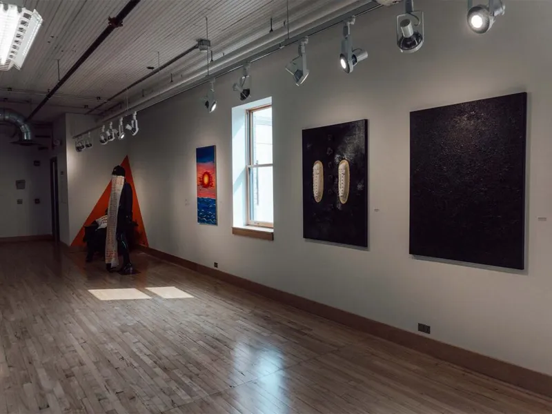 Gallery room with wood floors, track lighting, dark canvases, and a figure standing near an orange triangular installation