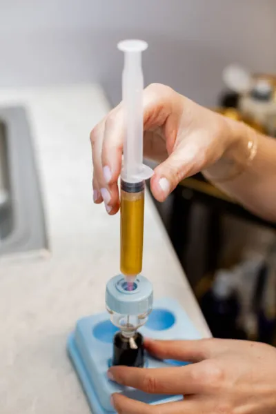 Hands drawing amber-colored liquid into a syringe from a small vial in a lab tray