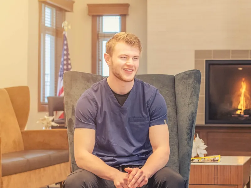 Smiling young man in blue scrubs seated in a gray wingback chair near a lit fireplace