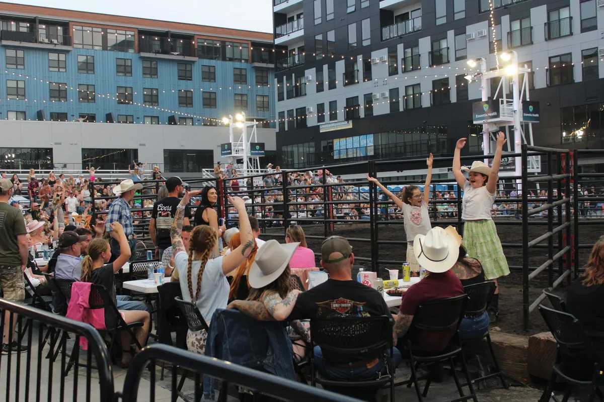 Crowd in cowboy hats cheers at an outdoor rodeo arena with string lights and urban buildings at dusk