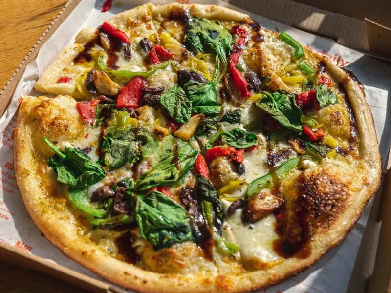 Veggie pizza with spinach, red peppers, olives, and balsamic drizzle in a takeout box