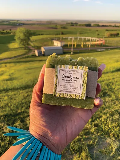 Hand holding a green Sweetgrass handmade soap bar above rolling farmland
