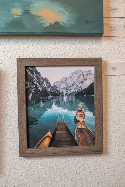 Framed painting of wooden boats on a turquoise mountain lake displayed on a gallery wall