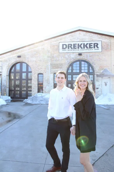 Couple smiling and holding hands outside Drekker Brewing Company in winter, woman displaying engagement ring