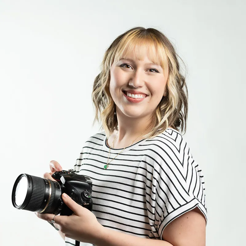 Smiling woman with wavy blonde hair holding a DSLR camera, wearing a striped top