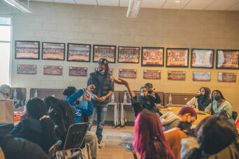 A man stands and gestures while speaking to a group of youth seated around tables in a community room