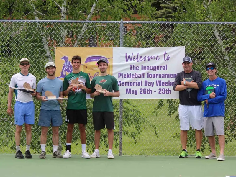 Six players pose with paddles and trophies in front of an inaugural pickleball tournament banner