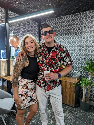 Two smiling people pose together inside a stylish studio with patterned wallpaper and copper balloons