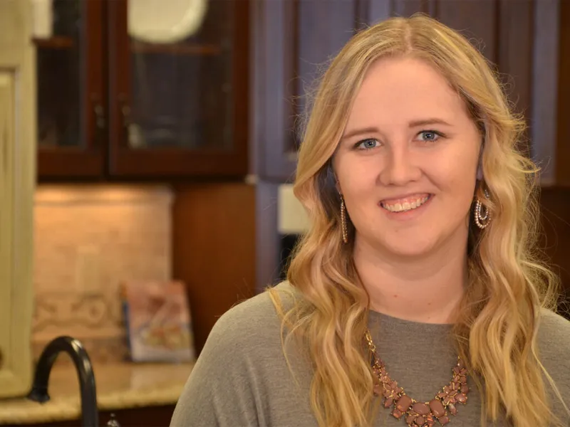 Smiling woman with wavy blonde hair and a statement necklace standing in a kitchen