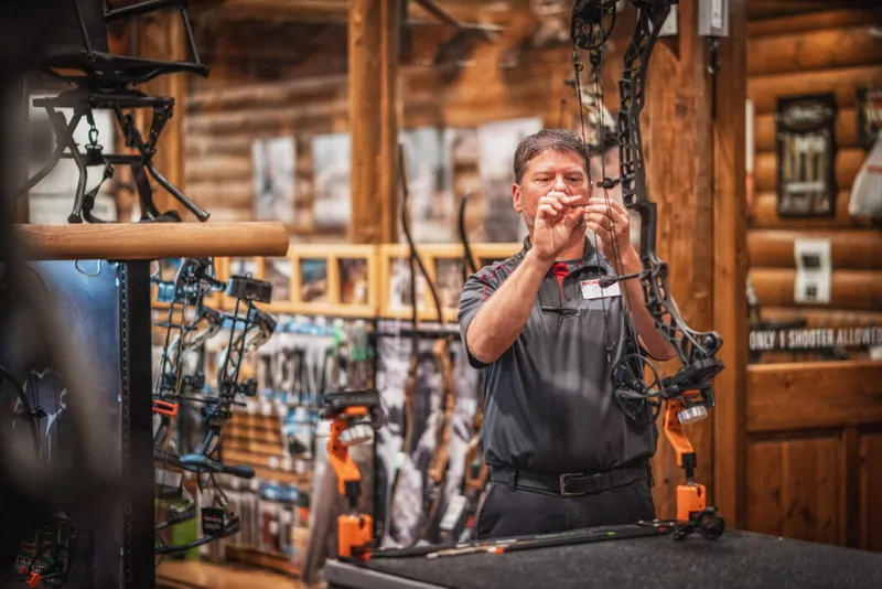 A store employee in a gray polo inspects a compound bow inside a rustic log-walled archery shop