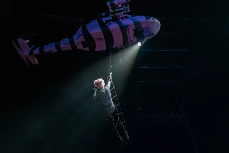 Pink-haired performer hanging from a ladder beneath a helicopter prop in a darkened arena