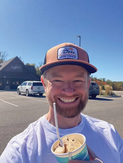 Smiling man in a trucker hat holding an ice cream cup outdoors on a sunny day