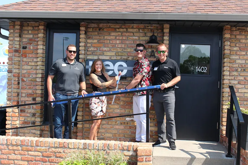 Four people cutting a blue ribbon outside a brick building with a neo sign