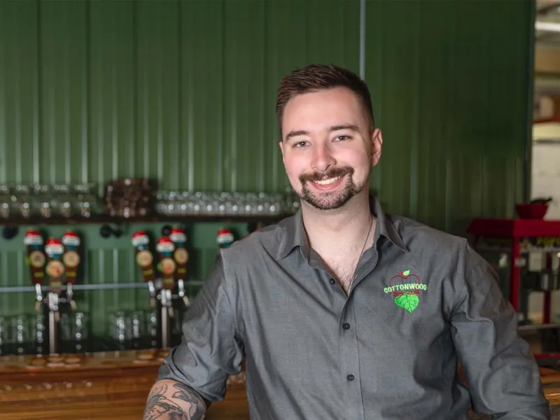 Smiling bartender in a gray Cottonwood logo shirt leaning on a wooden bar with beer taps behind him